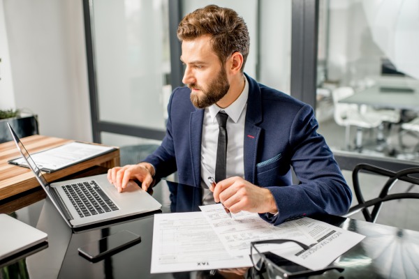 Mann im blauen Anzug arbeitet an einem Laptop, umgeben von Papieren und Smartphone, in einem modernen Büro mit Glaswänden.