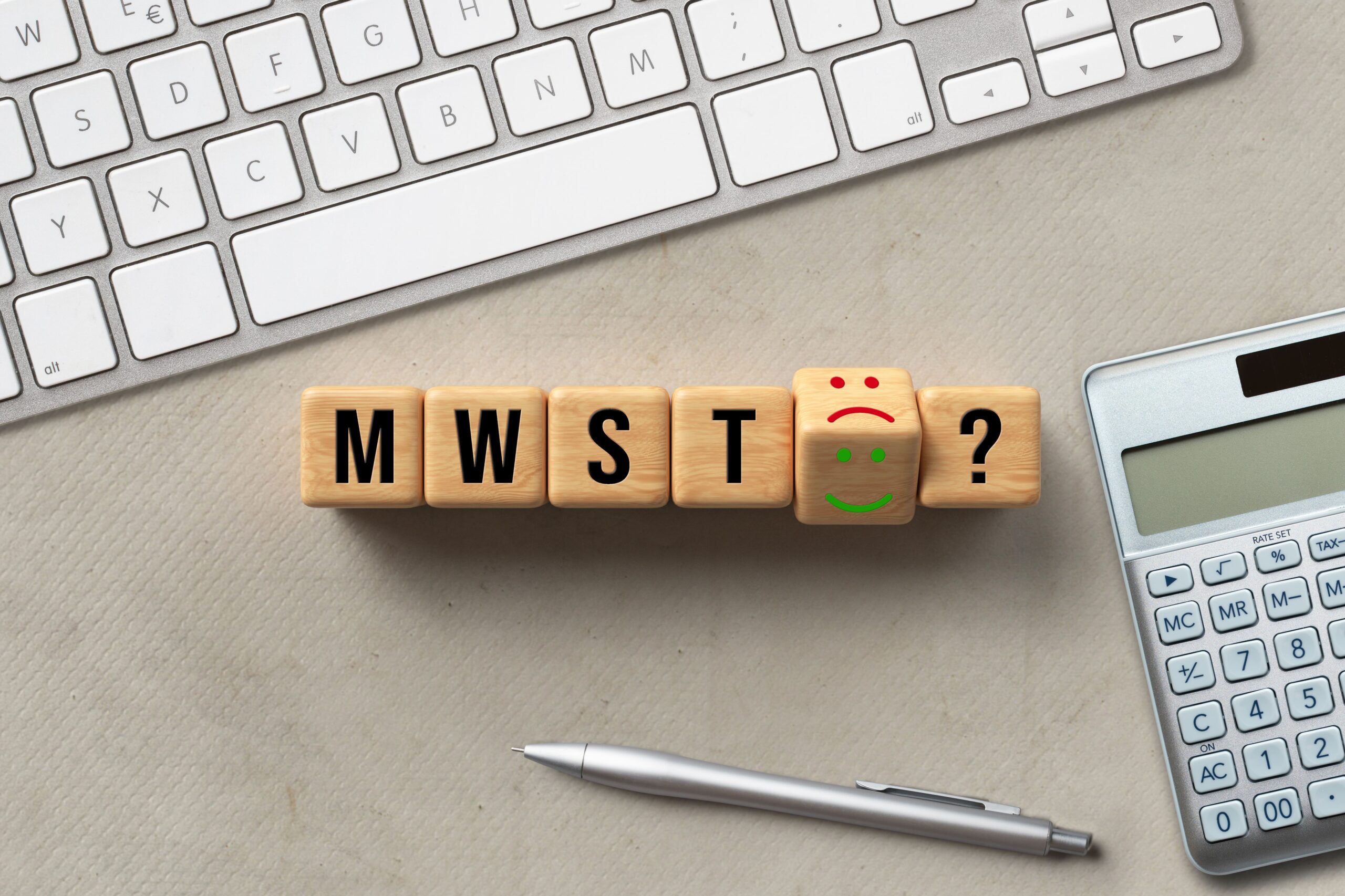 Holzklötze „MWST“ mit Smileys, silberner Stift, Taschenrechner und Tastatur symbolisieren Umsatzsteuer bei digitalen Produkten.