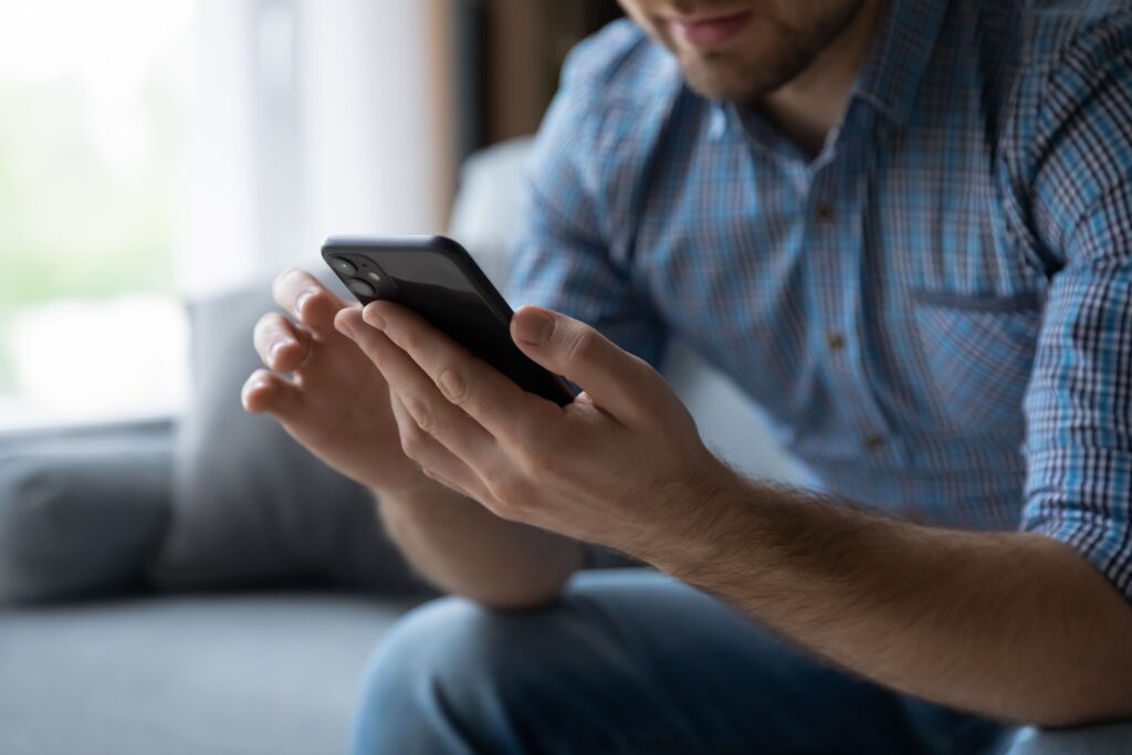 Ein Mann im karierten Hemd sitzt auf einem Sofa und konzentriert sich auf sein Smartphone, möglicherweise überprüft er digitale Umsatzsteuerinformationen.