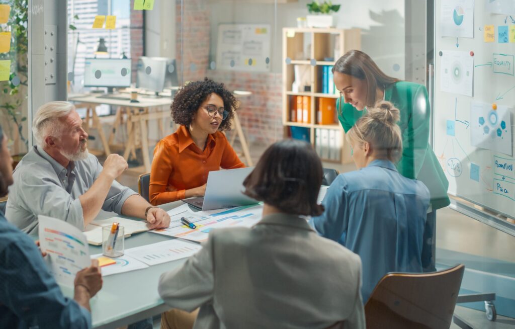 Diverse Gruppe in modernem Büro bei Meeting, nutzen Qualifizierungschancengesetz zur Weiterbildung; Person präsentiert, andere hören zu und machen Notizen.